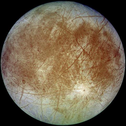 Image of Europa, Jupiter's moon, with a smooth, icy surface crisscrossed by dark brownish cracks and streaks, indicating possible subsurface oceans