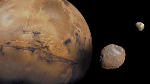 Image of Mars with its two moons, Phobos and Deimos, floating in space. The planet's surface shows detailed features like Valles Marineris, with the moons appearing as smaller, irregularly shaped objects in the foreground