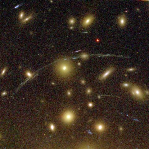 An image that shows many bright galaxies against a dark, fuzzy background. Across the centre, there is a large curved arc of light, with a few smaller arcs beneath this.