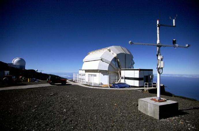 In the foreground there is a large, white aerial. Behind this, on the edge of a mountaintop, is a white telescope ensure with a clamshell roof. Another enclosure is in the distance on the left, behind a small slope of a hill. To the image right, a few clouds are below the mountain. The sky is bright blue in colour, though darker towards the top edges of the image.