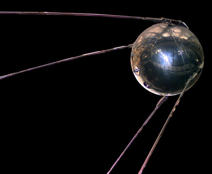 Shiny metallic sphere with four long antennas extending from its sides, against a dark background