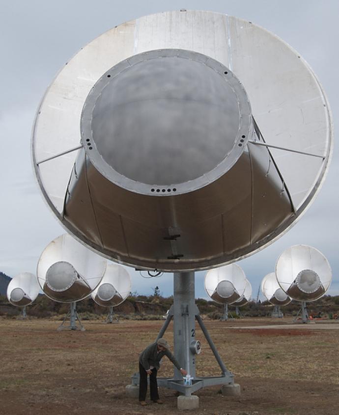 This image shows the Allen Telescope Array, a group of large, silver dish antennas used by the SETI Institute to search for extraterrestrial signals. One person is seen near the base of the foreground antenna, illustrating the massive size of the equipment