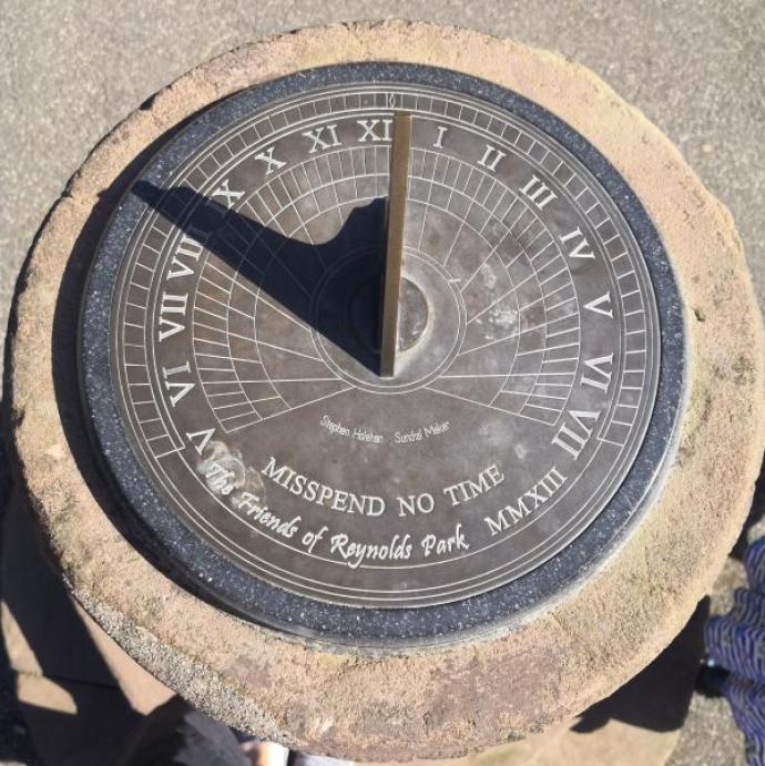 "A sundial with Roman numerals marking the hours and the words 'MISSPEND NO TIME' inscribed at the bottom. It also includes the text 'The Friends of Reynolds Park MMXIII' and the names 'Stephen Hales' and 'Sundial Maker.' The sundial is mounted on a stone pedestal, casting a shadow indicating the time