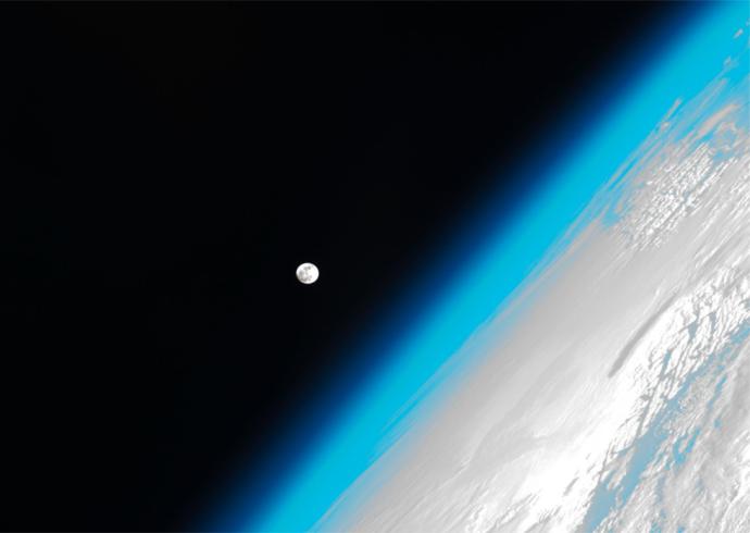 An image of Earth's atmosphere taken from space, with a clear gradient from the bright blue atmosphere to the blackness of space. The Moon is visible in the distance, with part of Earth's cloud-covered surface shown along the right edge of the image