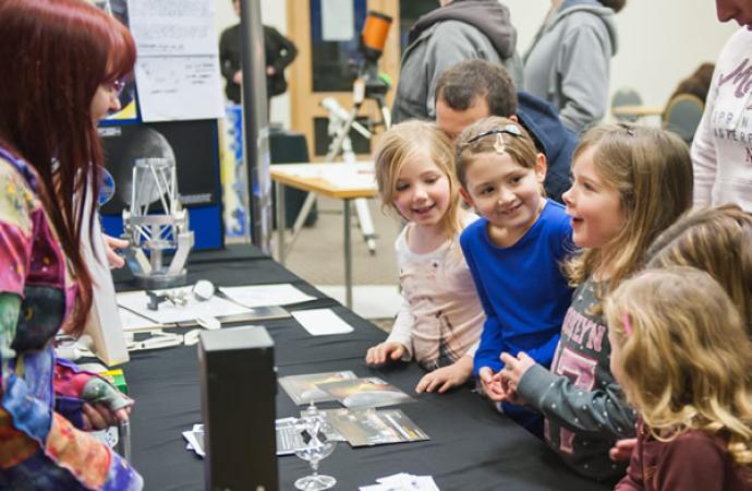 A group of young children excitedly engaging with a presenter at a science exhibit, with various materials and models displayed on the table