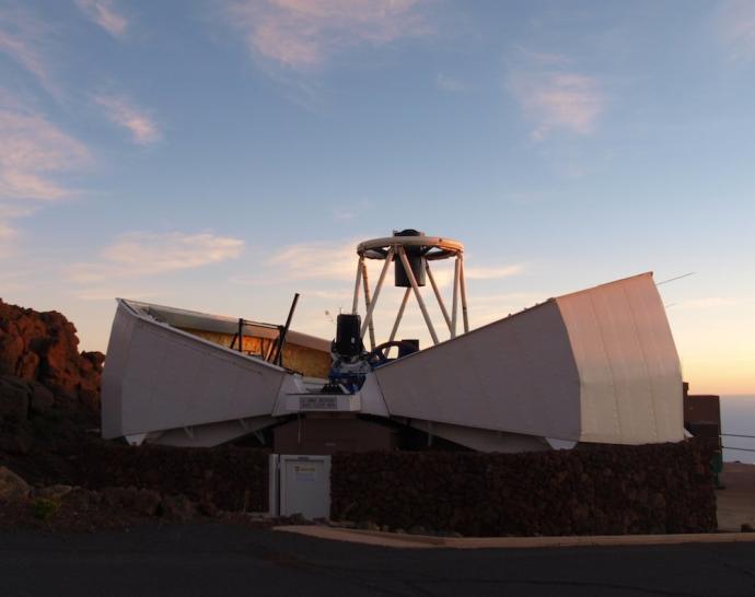 A clamshell telescope dome that is open. Inside, a telescope points straight up at the sky. The sky is light blue with a few thin clouds in places.