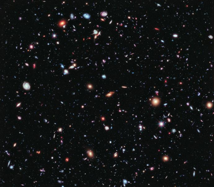 Many, many galaxies against a black background. Some are tiny dots, and they range in size all the way up to small spheres or swirls. They have different shapes, some more spiraled than others, whilst some appear like smudged lines. They range in colour from red, orange, white, green, and blue.