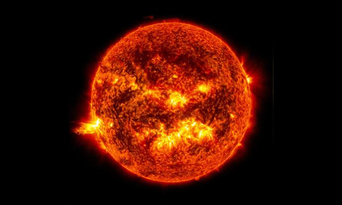 The sun against a black background. It is a dark orange sphere with some brighter yellow patches as well as some dark brown/black patches.